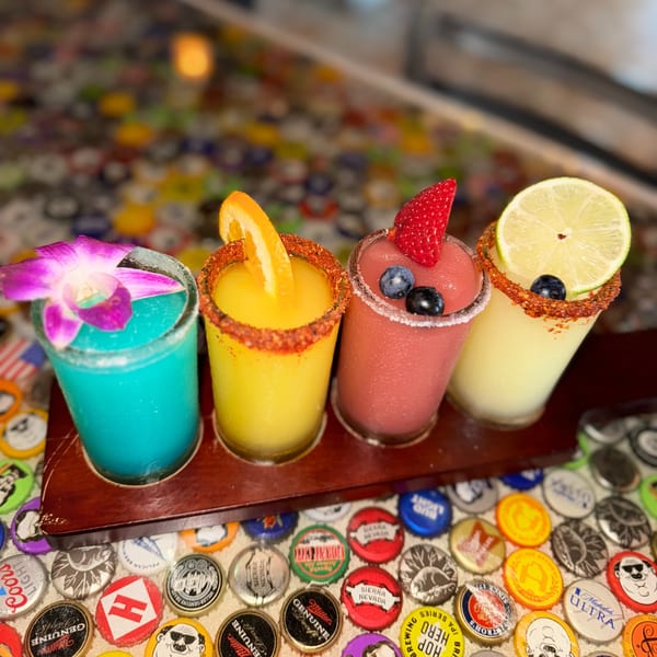 Four colorful frozen margaritas in a flight, garnished with fruit and a flower, on a bottle cap table.