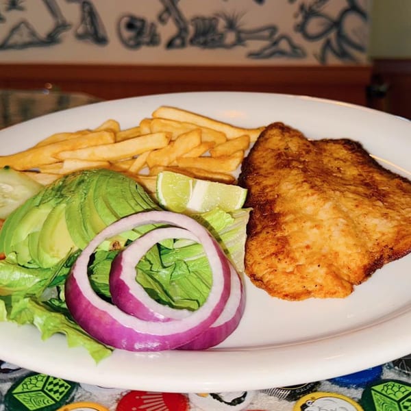 Fried fish fillet with french fries, avocado, red onion, lettuce, and lime on a white plate.