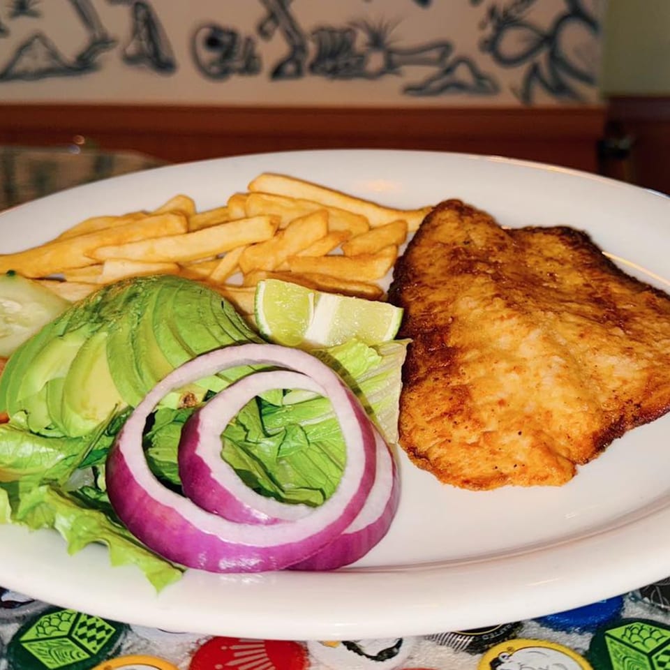 Fried fish fillet with french fries, avocado, red onion, lettuce, and lime on a white plate.